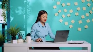 Asian administrative assistant works on a laptop at her desk. Office setting with sticky notes on the wall.