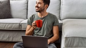 Adult learning mathematics online. Man with laptop and coffee cup smiles, seated on floor.
