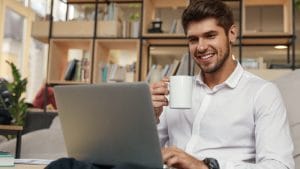 Smiling man drinking his morning coffee and working on his laptop.