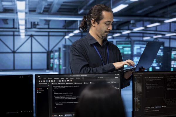 Network admin to site reliability engineer. IT professional with laptop in data center, coding on monitors.