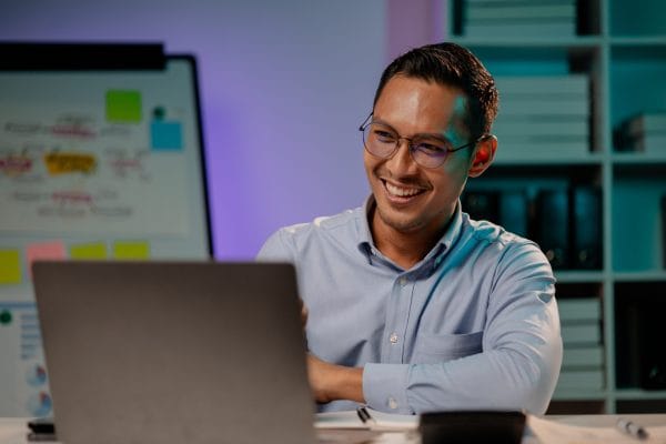 Smiling man using no/low code machine learning on laptop. Whiteboard with sticky notes in background.