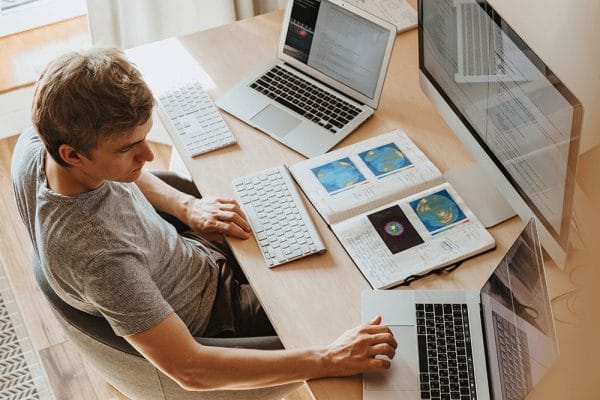 CompTIA Bundle: Man at desk with laptops, keyboard, monitor, and notebook.