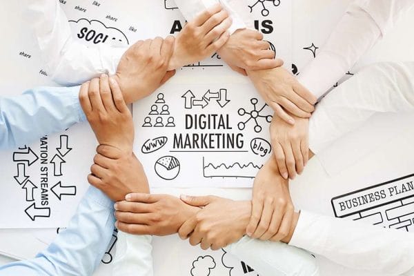 Teamwork in digital marketing. Hands linked around Digital Marketing sketches, social media, analysis, & business plan.