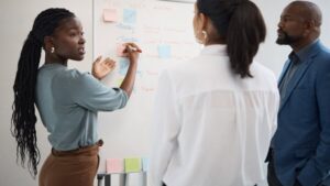 Professional woman with braided hair places a sticky note on a whiteboard while two colleagues observe in an office.