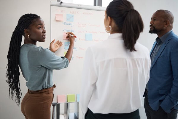 Professional woman with braided hair places a sticky note on a whiteboard while two colleagues observe in an office.